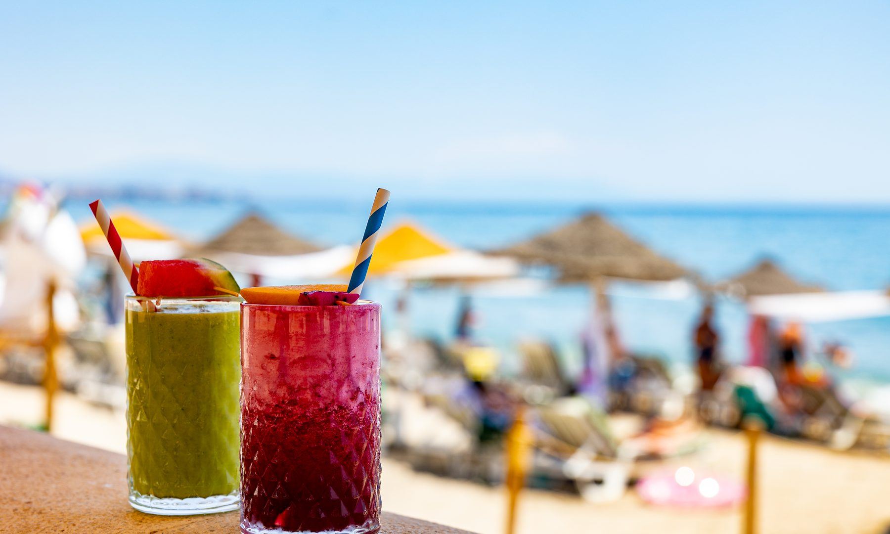 A green juice and a berry juice on the sea wall, straw parasols and the Mediterranean behind.